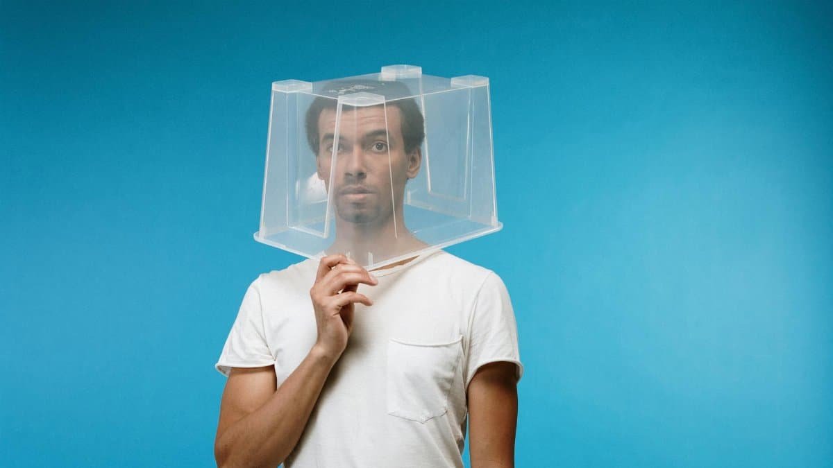 A man humorously uses a plastic box as a DIY protective face shield against a blue backdrop.