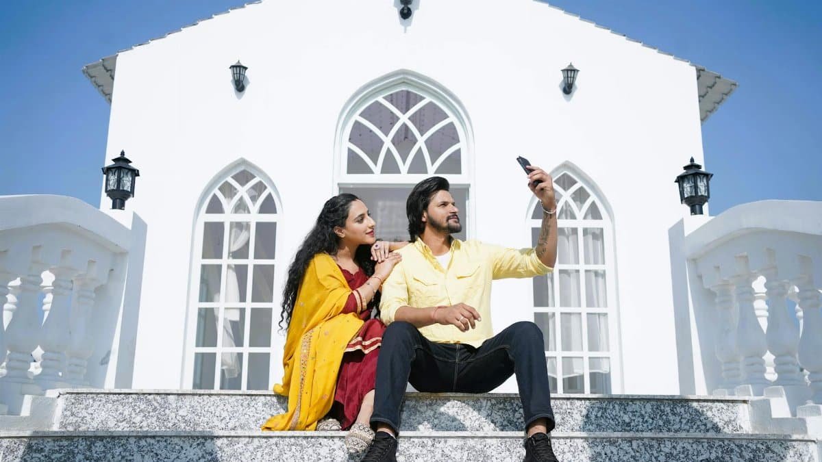 A couple sitting on steps, taking a selfie in front of a modern house with arched windows.