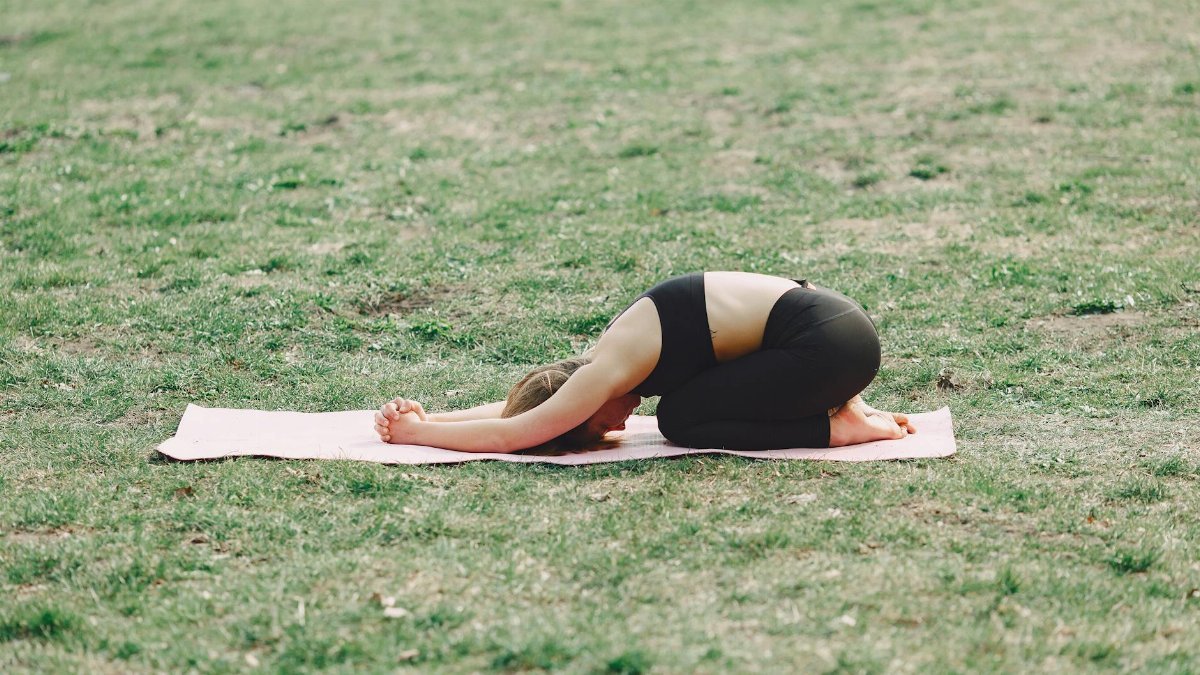 A woman practicing yoga outdoors in a park, in a peaceful child's pose on a mat.