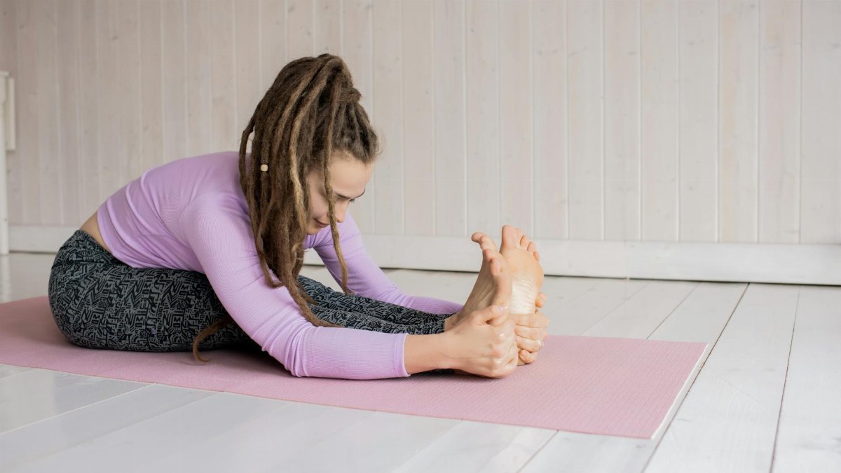 A woman stretching in a seated forward bend yoga pose on a pink mat indoors.