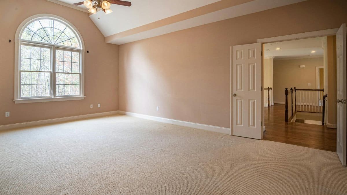 Bright empty room with beige walls, carpet flooring, and a large window showcasing modern architecture.
