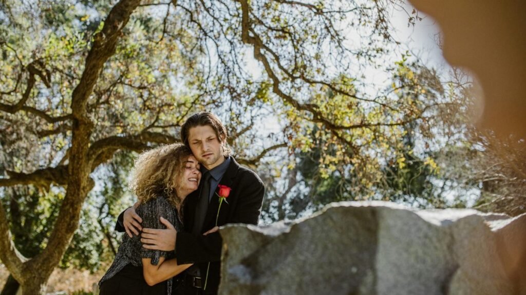 A couple embraces in grief, surrounded by nature in a sunny cemetery setting.