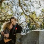A couple embraces in grief, surrounded by nature in a sunny cemetery setting.
