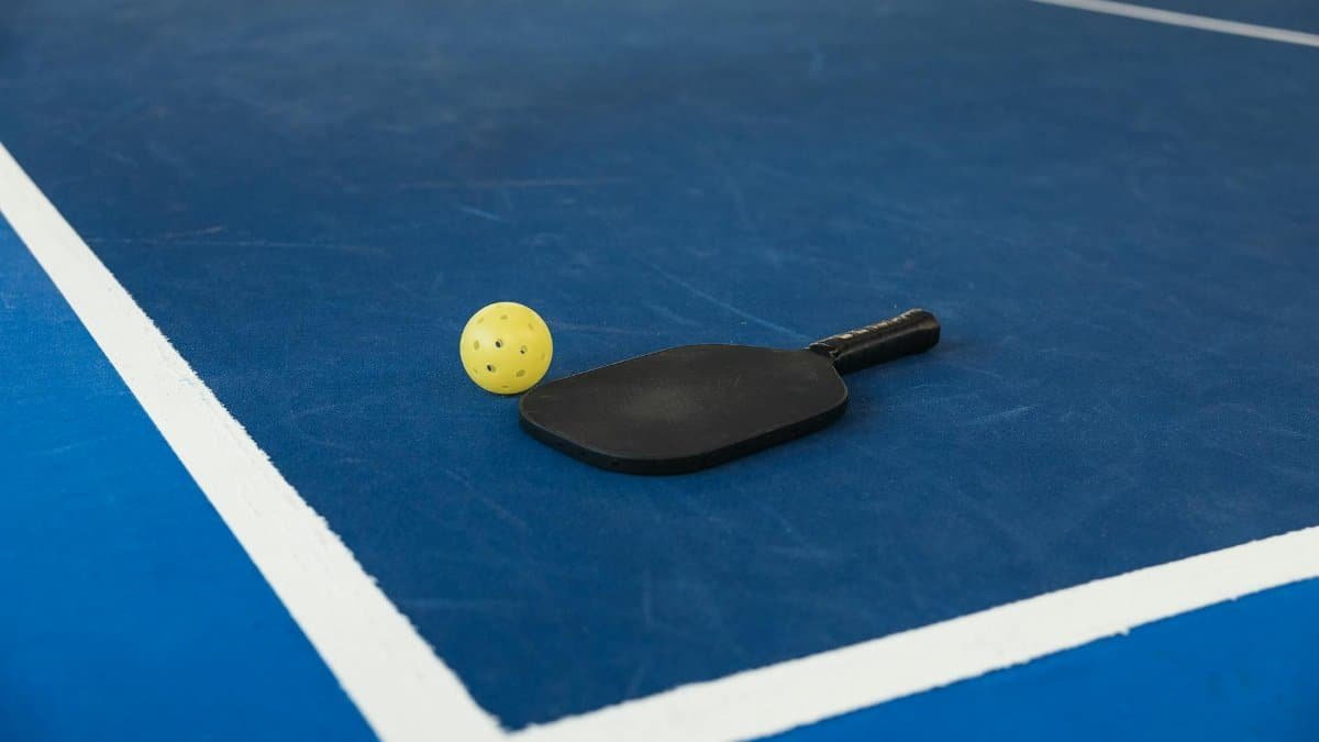 A pickleball paddle and yellow ball resting on a blue pickleball court with white lines.