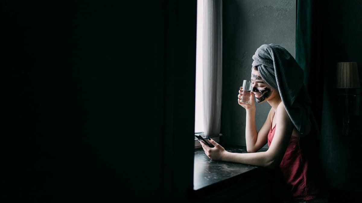 Woman with facial mask and towel turban enjoying skincare routine indoors, holding a glass of water.