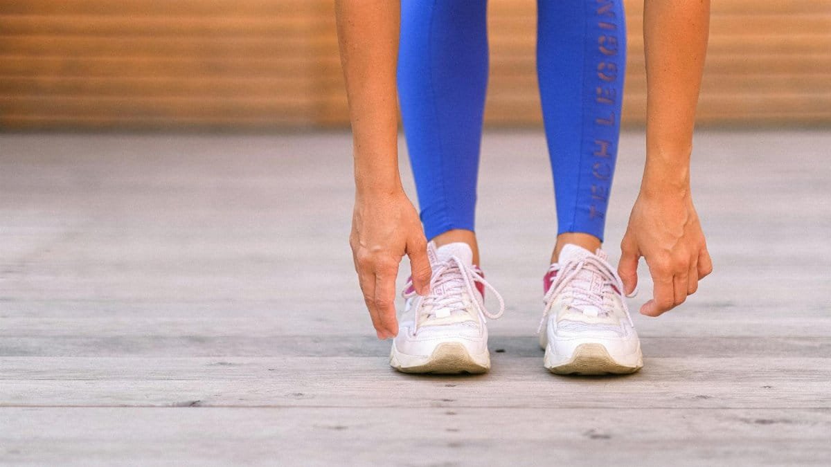 Crop anonymous sportswoman in blue leggings and sneakers stretching body and bending forward while warming up before exercising in fitness studio