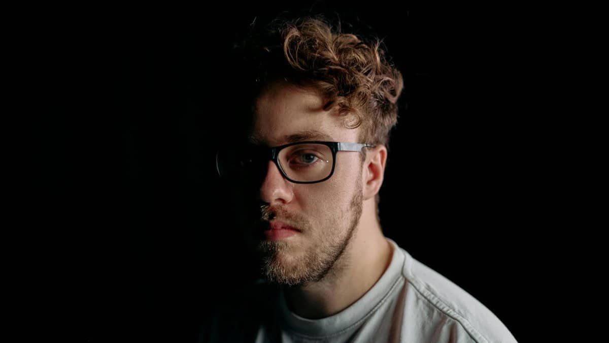 A dramatic half-face portrait of a man wearing glasses, captured with soft focus and low-key lighting.