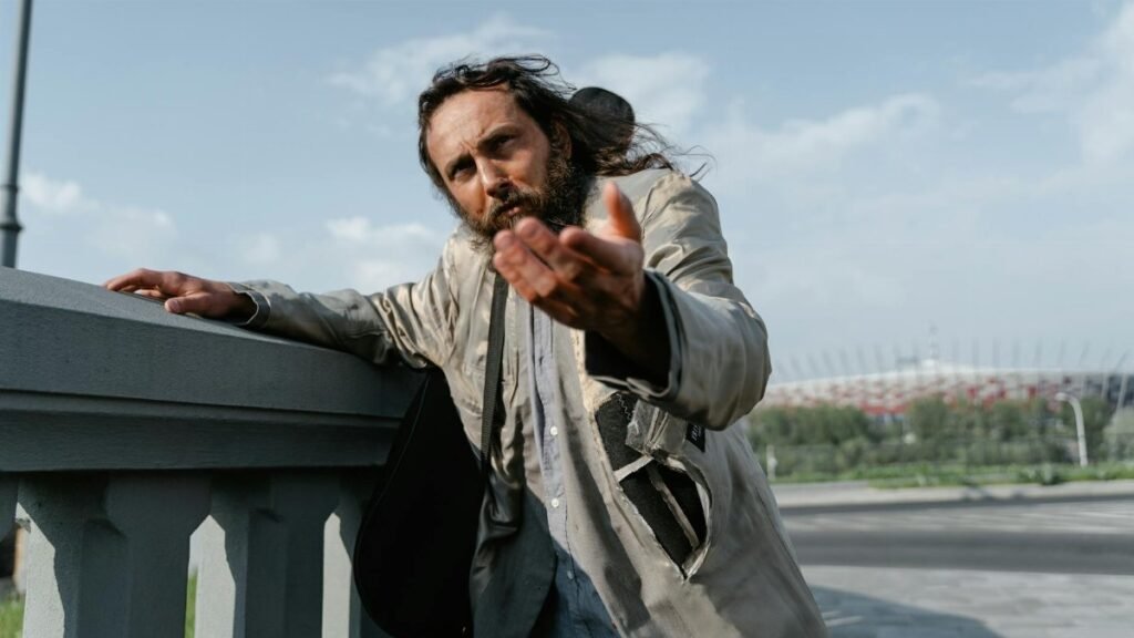 A homeless man in tattered clothing extends his hand, pleading for help on a sunny day.
