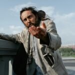 A homeless man in tattered clothing extends his hand, pleading for help on a sunny day.