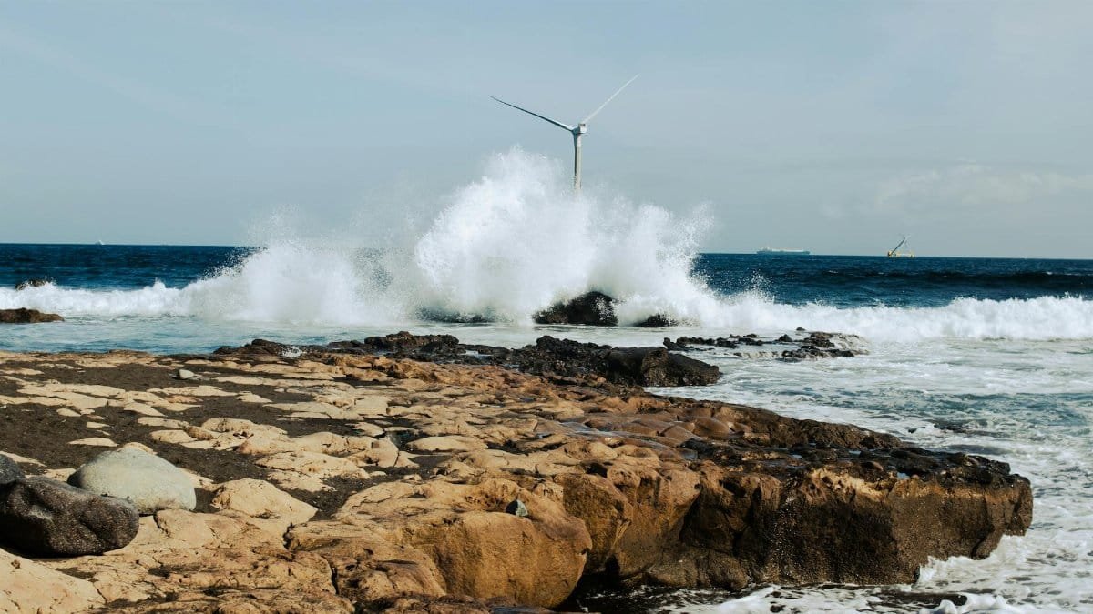 A stunning view of ocean waves crashing on rocks with a wind turbine in the background, highlighting renewable energy.