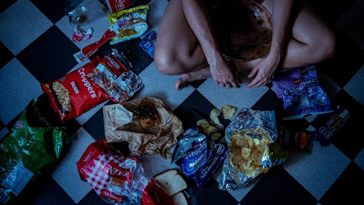 Man sitting surrounded by assorted snacks and packaging on a checkered floor, depicting a binge-eating scene.