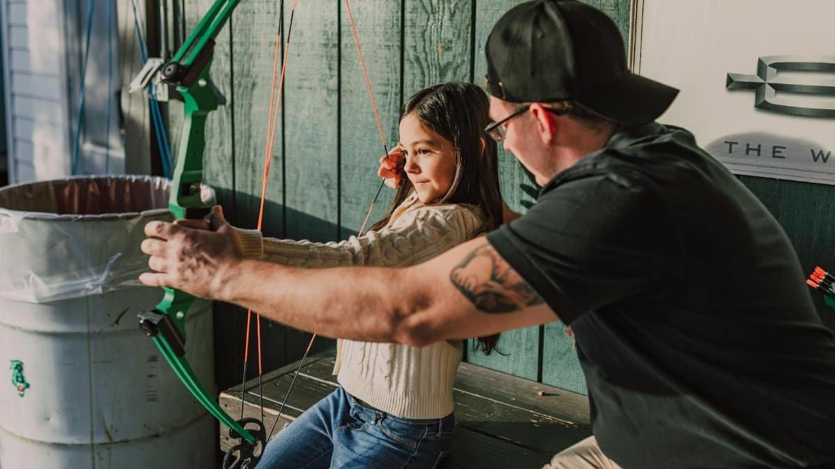Young girl being taught archery by a male instructor outdoors. Archery training session.
