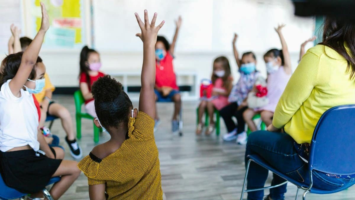 Children in a preschool classroom raise hands during a lesson on a school day, emphasizing diversity and learning.