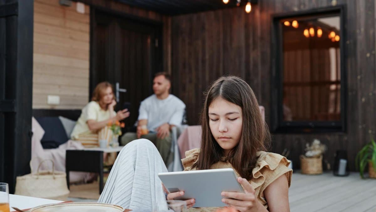 Teen girl focused on a tablet while sitting on a cozy outdoor patio with family in the background.