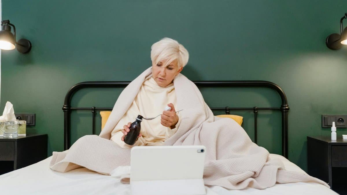 A senior woman sits on her bed using telehealth services during a pandemic, engaging with a health professional online.