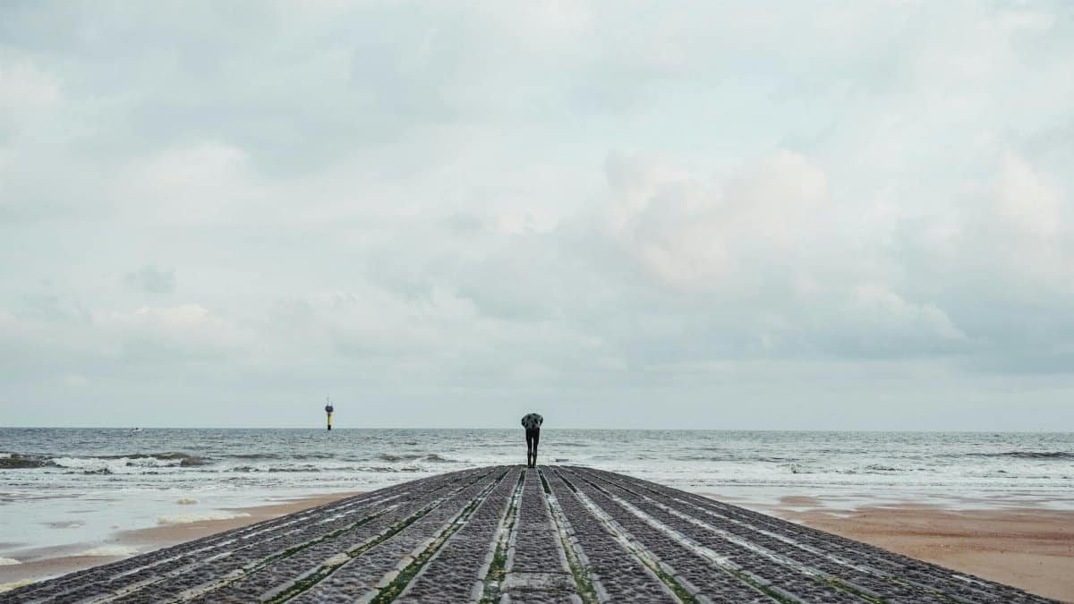 A lone person stands on a pier at Knokke-Heist beach, facing the sea under a cloudy sky.