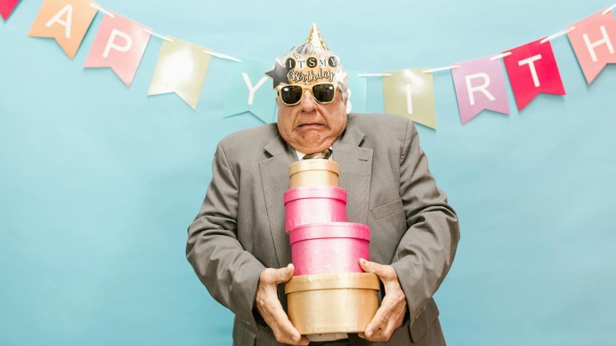 Senior man in party hat holding gift boxes against a colorful birthday banner.