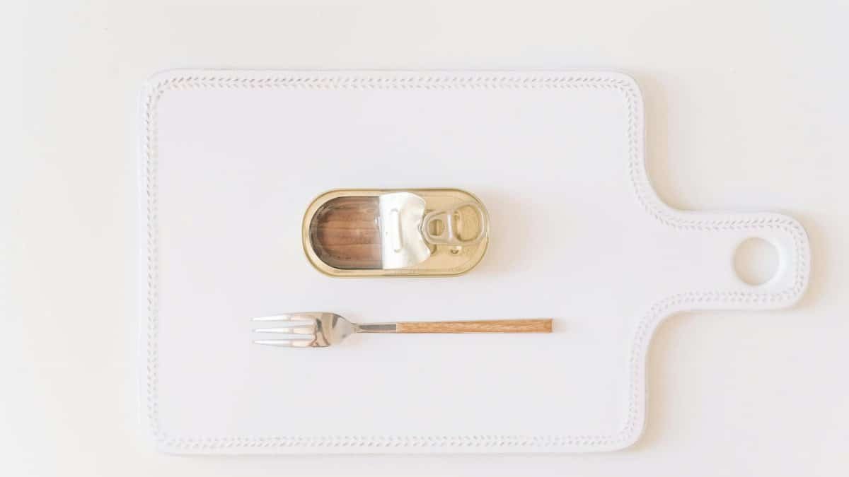 A minimalist composition featuring an open canned fish with a fork on a decorative white cutting board.