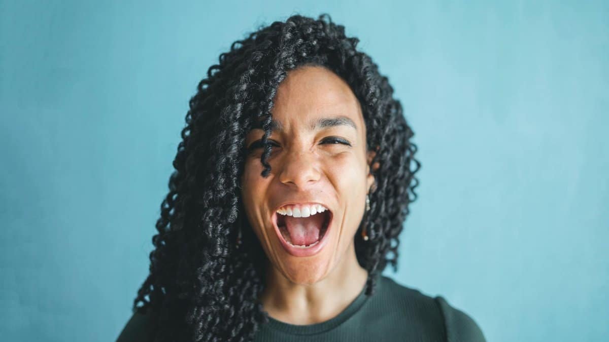 Portrait of a joyful young woman with curly hair, expressing excitement and shouting indoors.