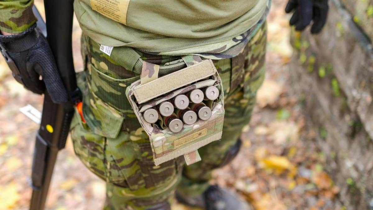 Close-up of a soldier in camouflage uniform with ammunition and shotgun outdoors.