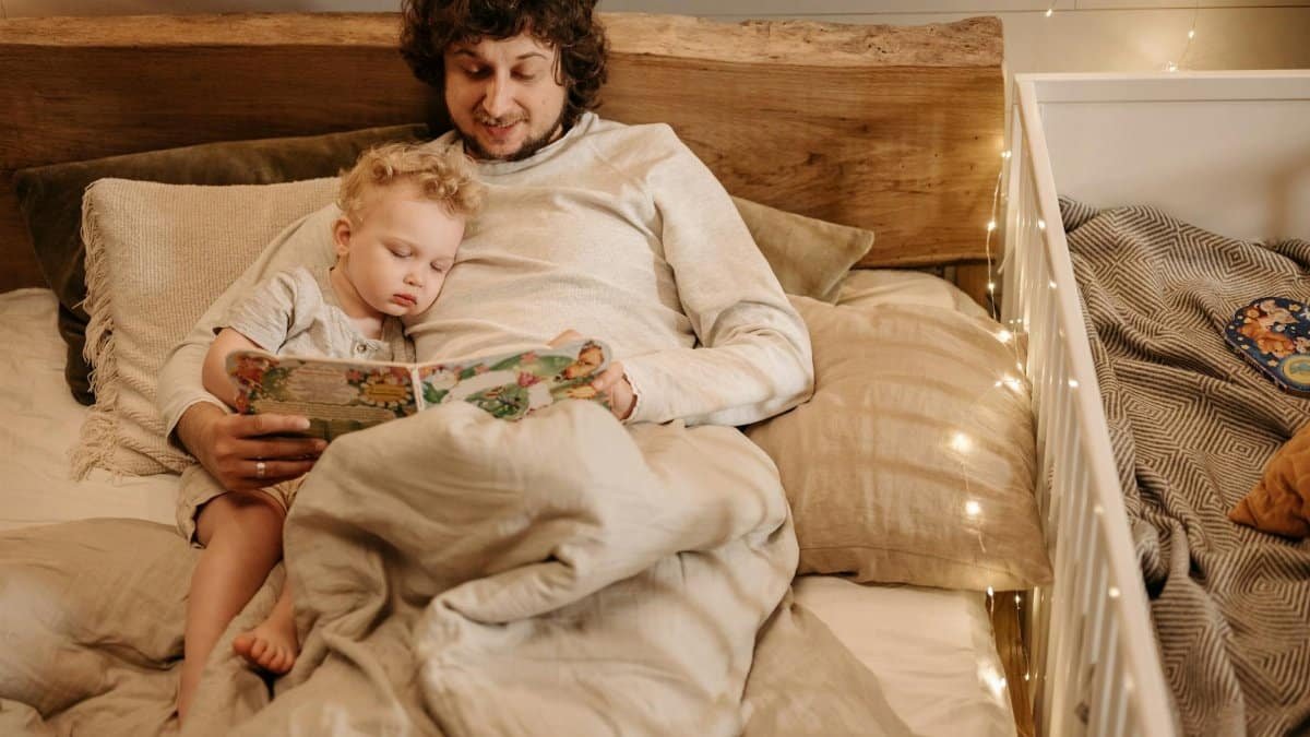 A father and child share a cozy bedtime story in bed, surrounded by warm lighting.