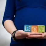 Pregnant woman in blue dress holds 'boy' blocks, symbolizing a baby boy announcement.