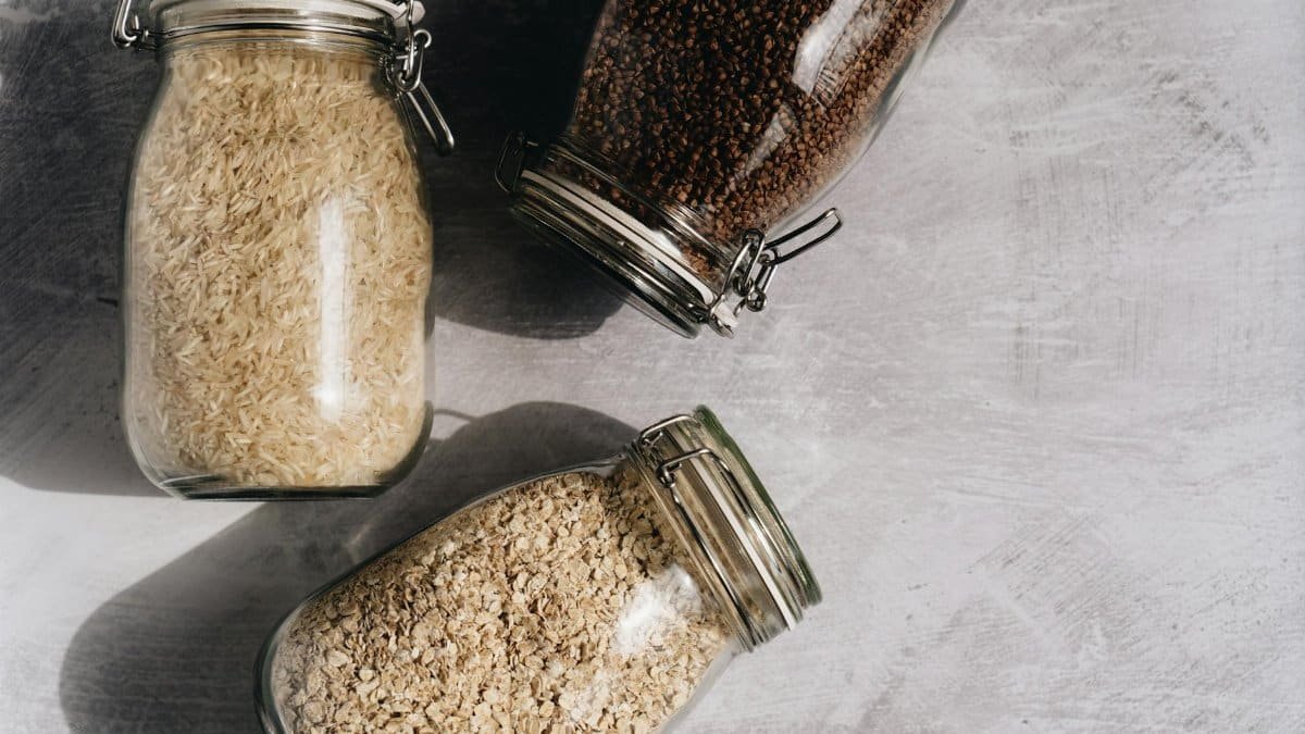 Top-down view of rice, oats, and buckwheat in glass jars on a textured surface.