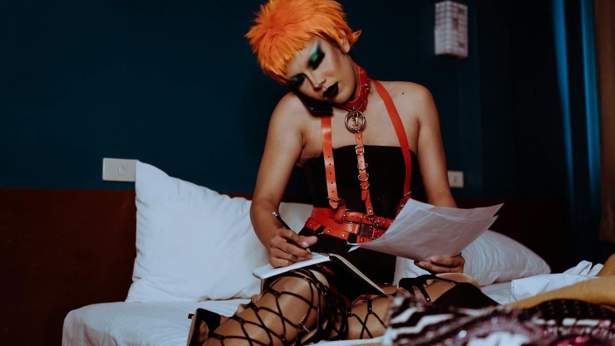 Androgynous individual with orange wig taking notes on bed during phone call.