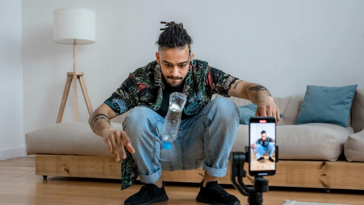 A young man focuses on a bottle flip challenge while filming content at home, capturing it on a smartphone.