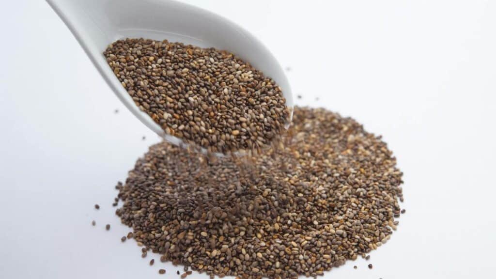 A close-up shot of chia seeds pouring from a white ceramic spoon onto a white surface.