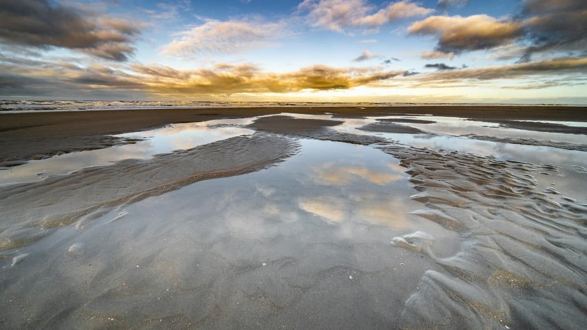 Serene beach scene with reflections in tide pools and a vibrant sunset sky.