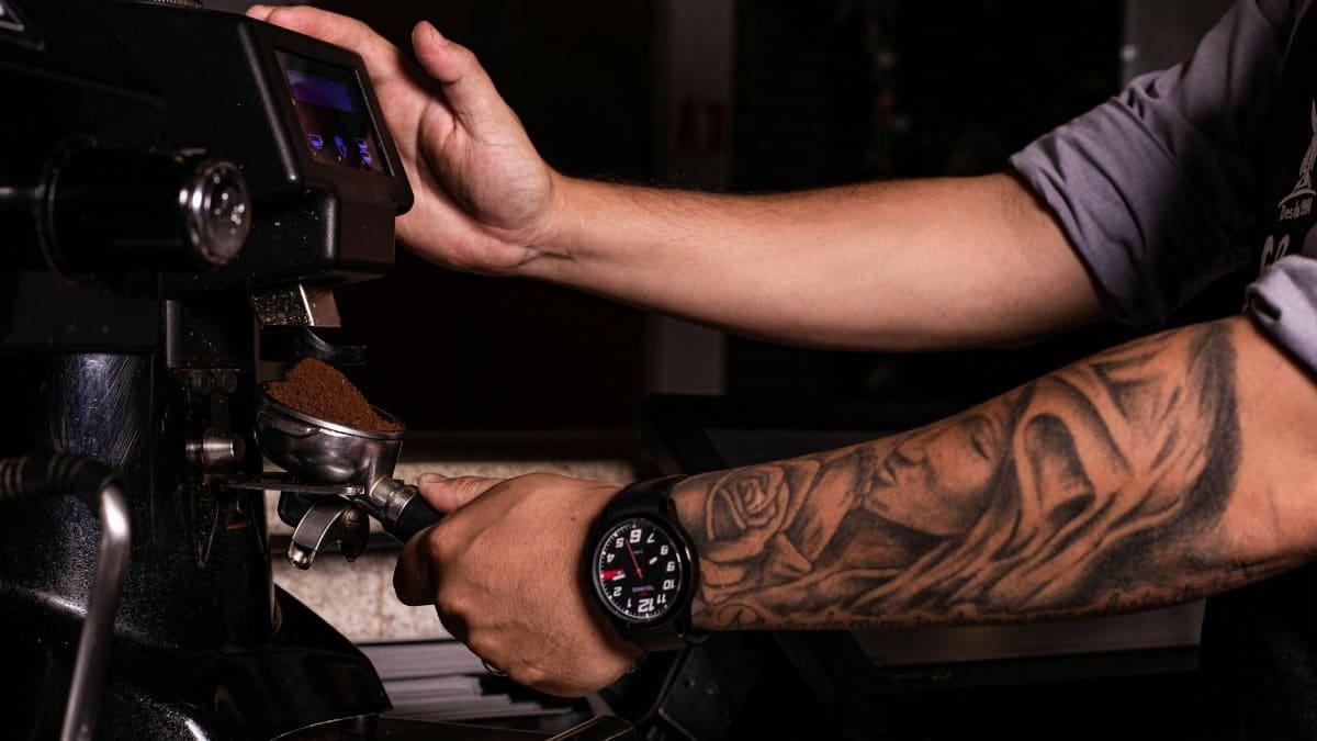 Close-up of a barista's tattooed arm using an espresso machine to grind coffee.