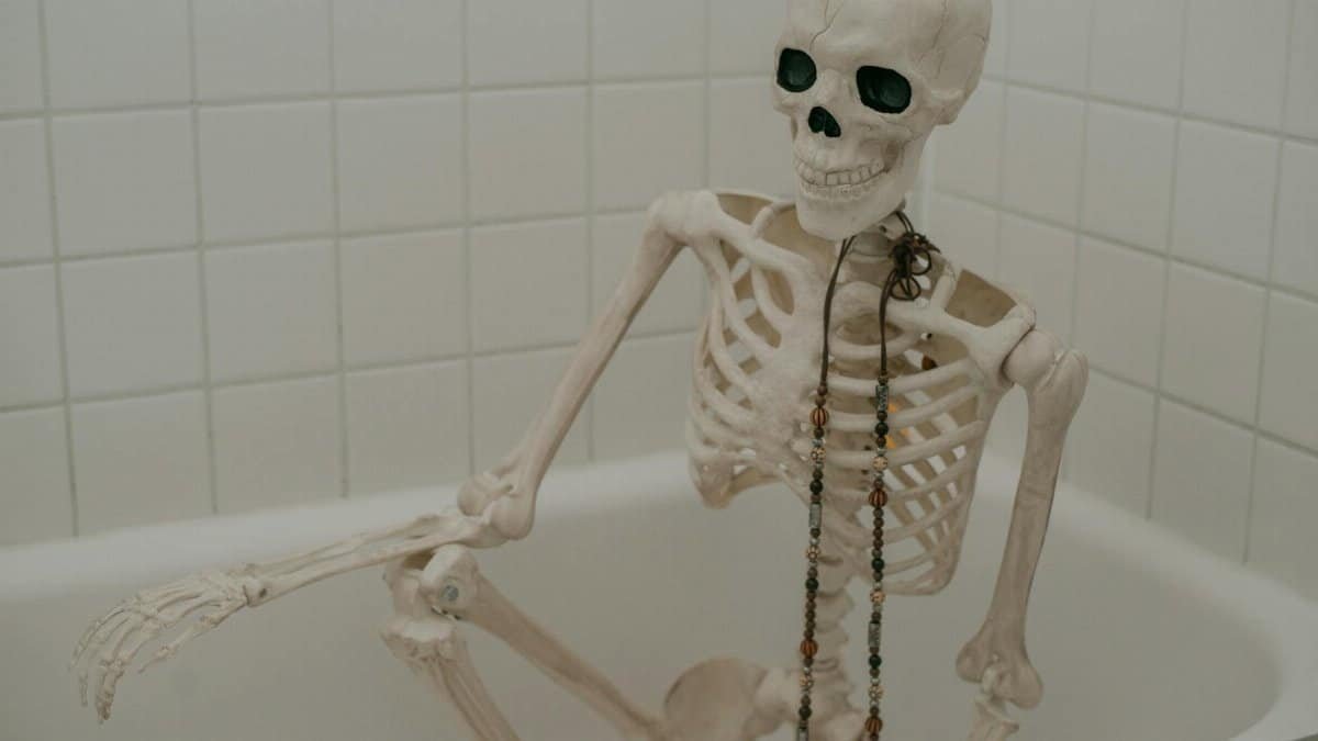 White skeleton with long chaplet sitting in bath without water in bathroom with white tiles on wall