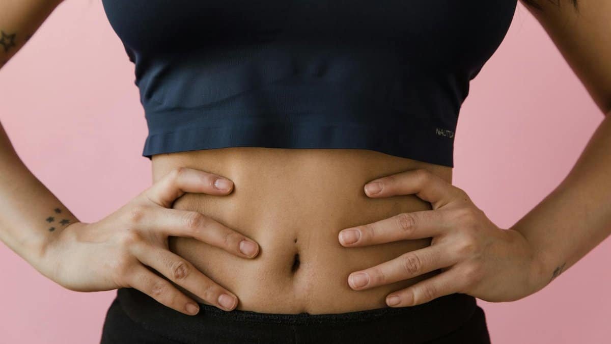 A close-up shot of a woman's toned stomach with hands on hips, against a pink background.