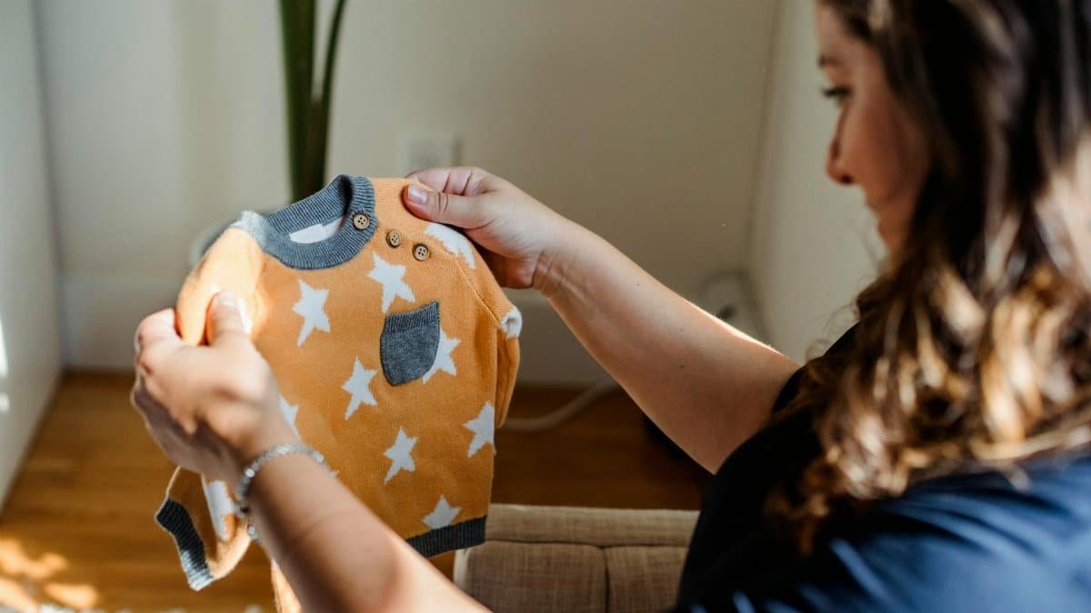 Side view of crop happy female choosing clothes for newborn baby while sitting on sofa at home