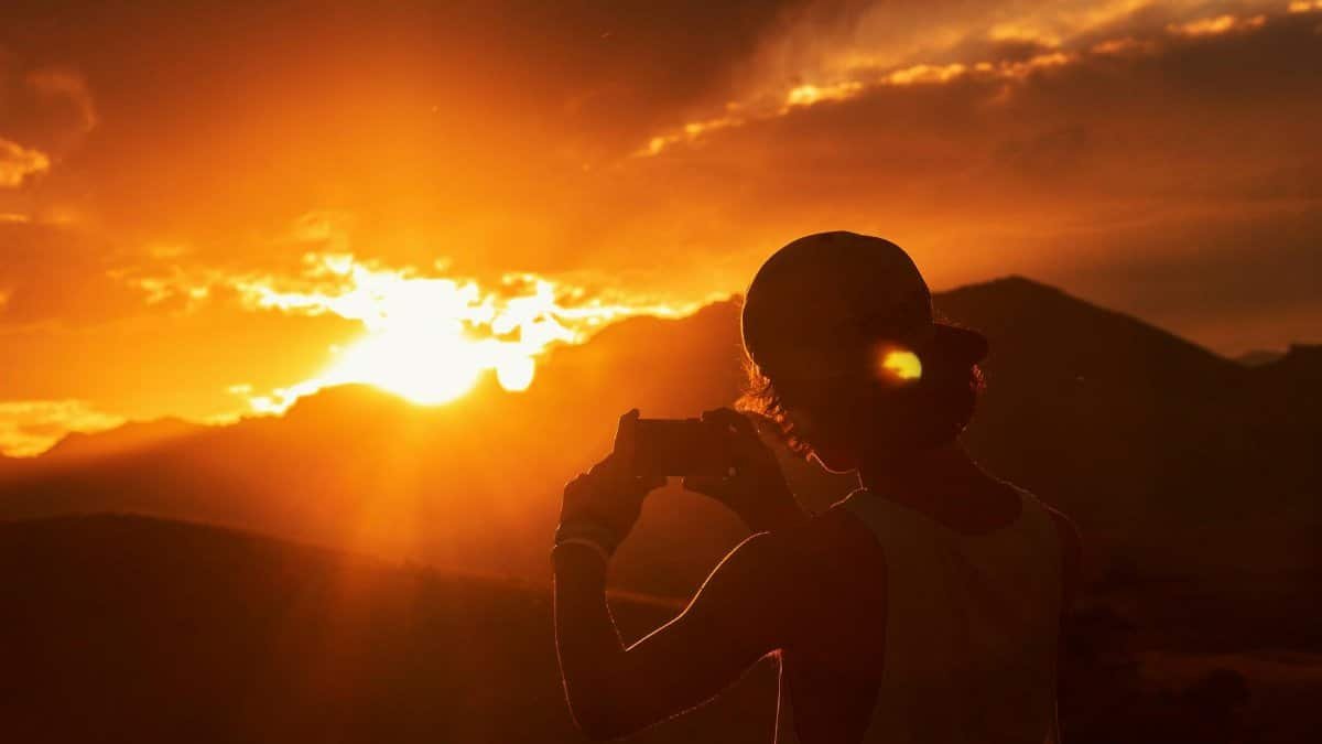 A silhouette of a person taking a photo against a dramatic sunset over mountains.