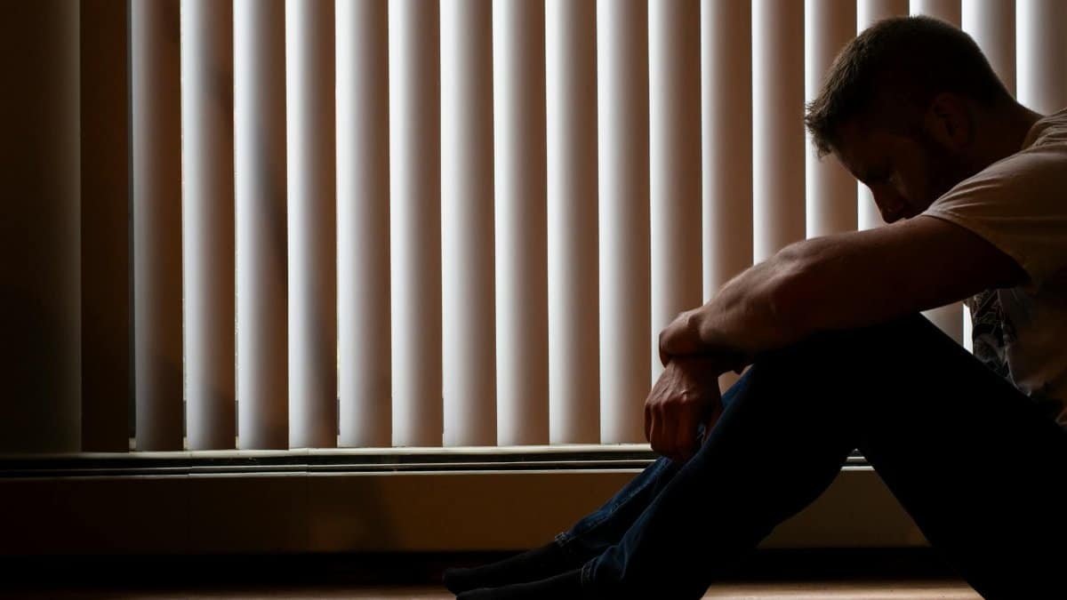 A man sits silhouetted against window blinds, conveying solitude and introspection.