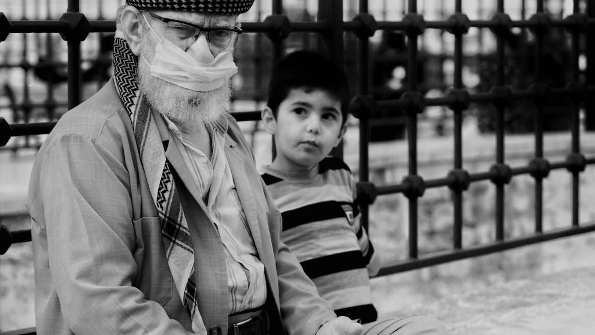 Black and white photo of an elderly man wearing a mask sitting with a boy outdoors, emphasizing generational connection.