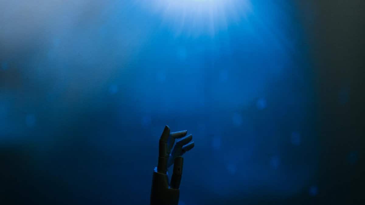 Silhouette of a robotic hand reaching towards glowing blue light in a futuristic setting.