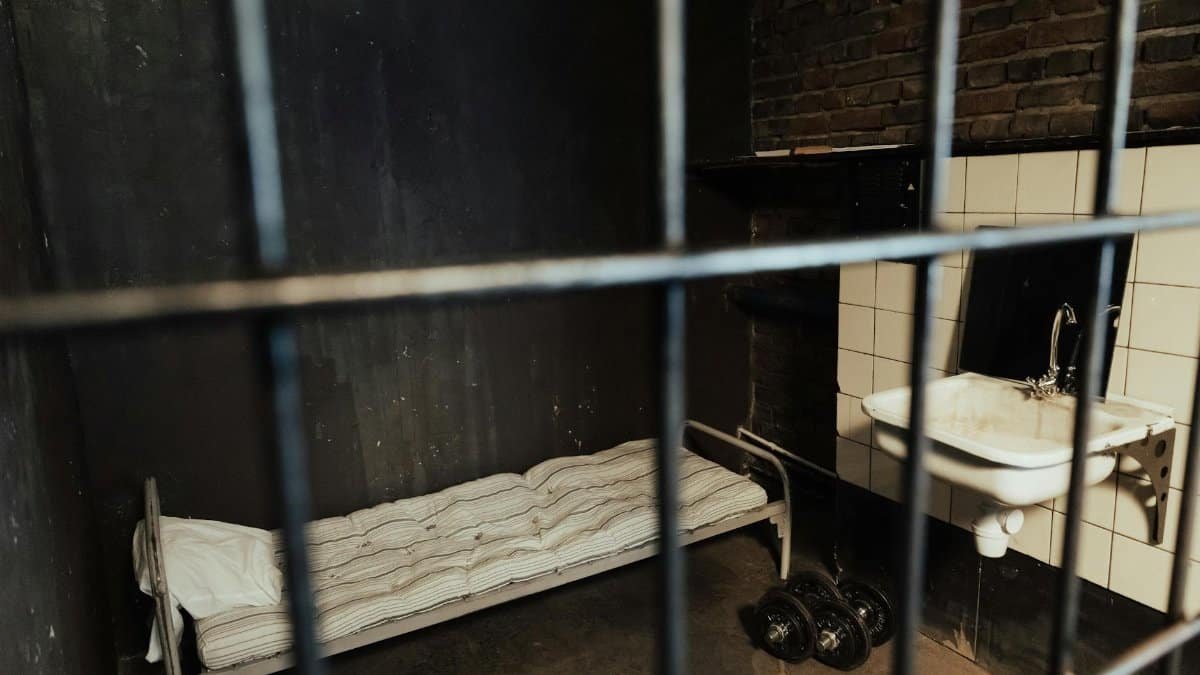 Moody prison cell interior featuring a metal bed, sink, and dumbbells behind bars.