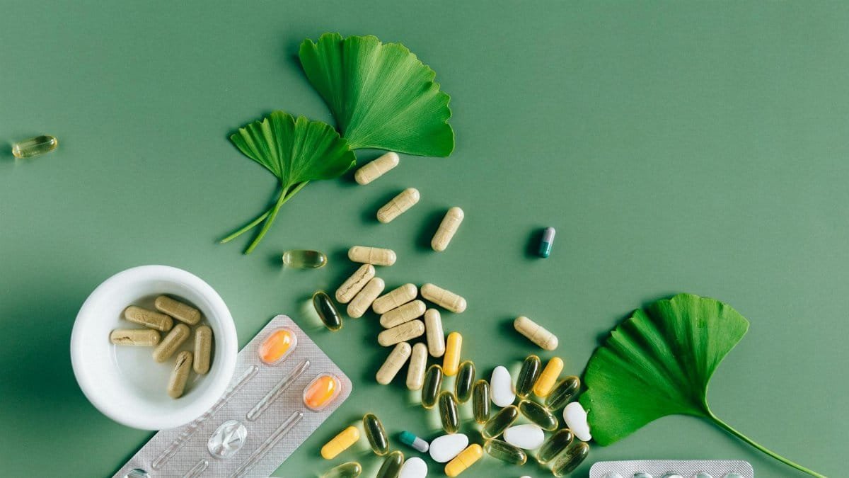 Flat lay of varied supplements and medicines with ginkgo leaves on green surface, showcasing a natural remedy concept.