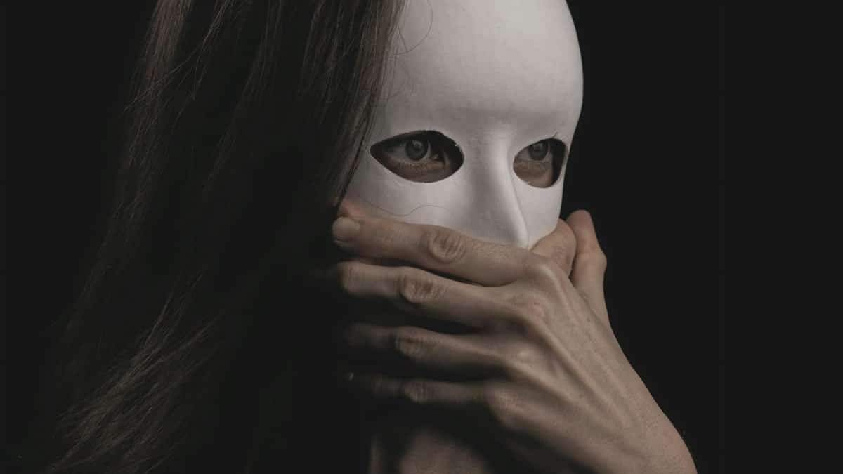 Close-up of a woman in a white mask, hands covering mouth, evoking mystery and drama.