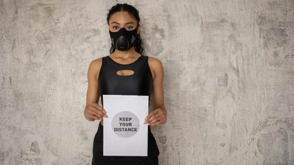 Young woman in sportswear wearing a mask and holding a 'keep your distance' sign against a neutral wall.