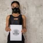 Young woman in sportswear wearing a mask and holding a 'keep your distance' sign against a neutral wall.