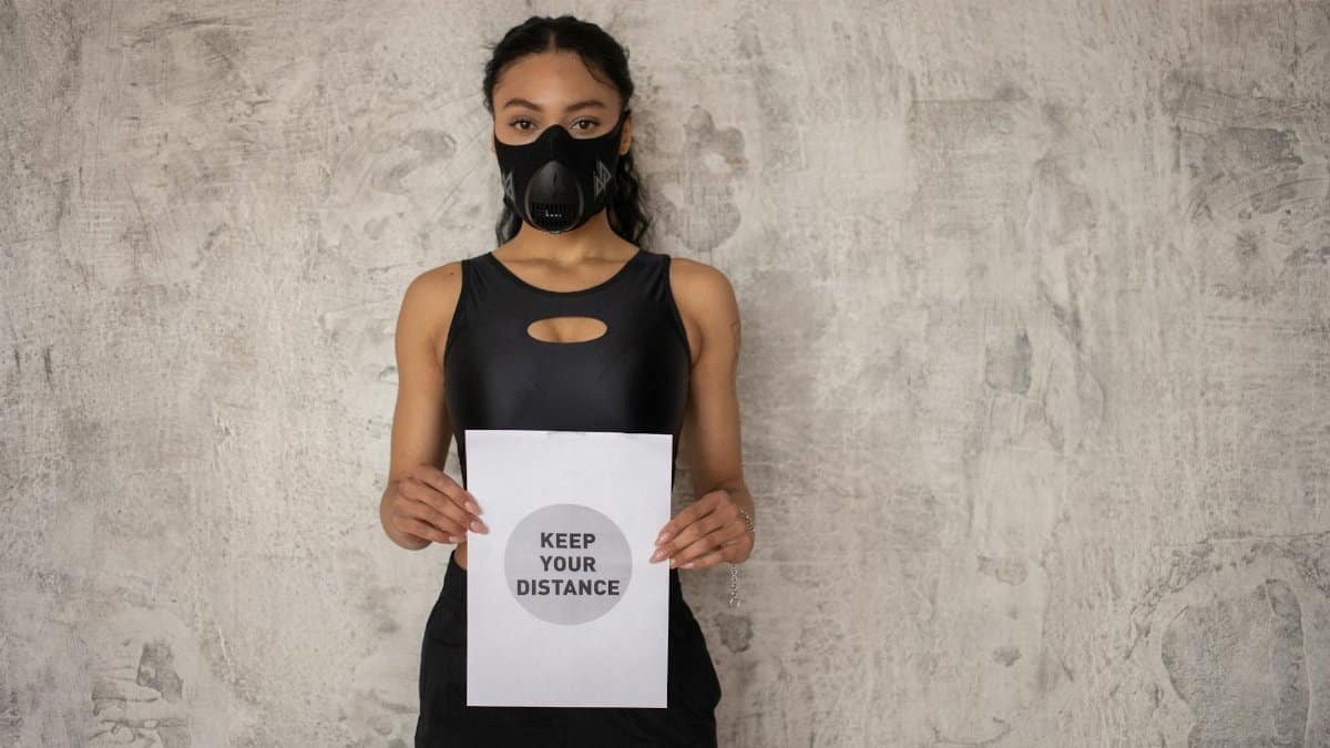 Young woman in sportswear wearing a mask and holding a 'keep your distance' sign against a neutral wall.
