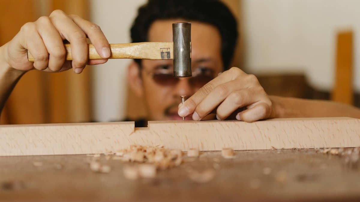A skilled carpenter focuses intensely on woodworking, using a hammer and nails in a bright indoor workshop.