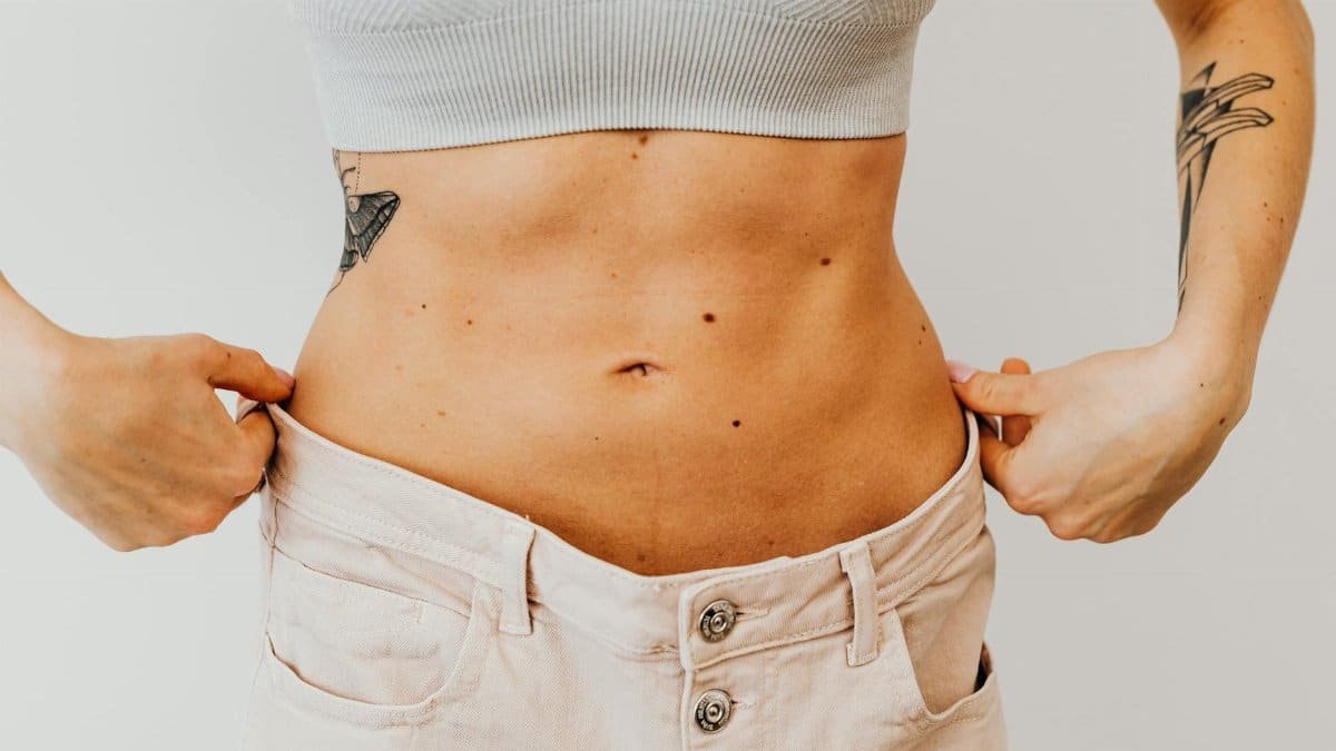A woman shows weight loss progress by holding loose pants around her waist, highlighting tattoos.