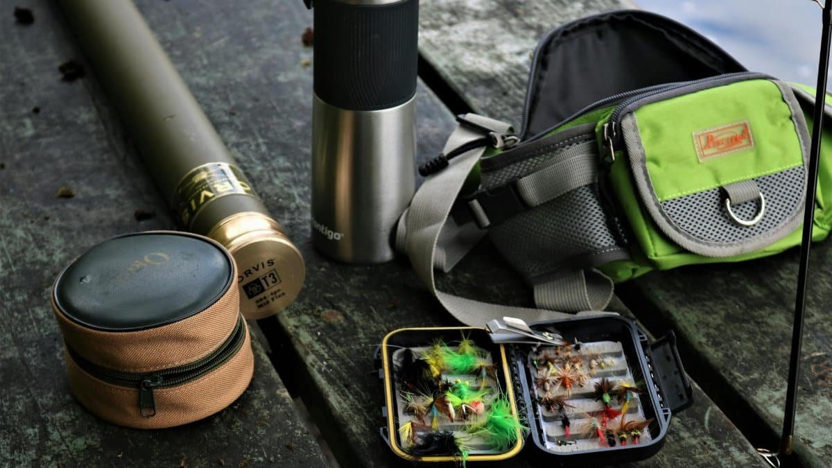 A detailed display of fishing equipment including fly box, backpack, and thermos, set outdoors.