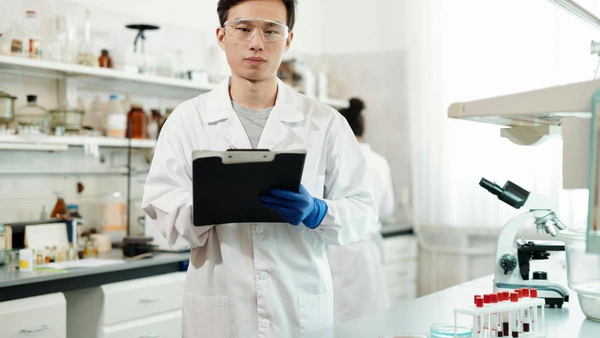 A young male scientist wearing a lab coat and safety glasses analyzes data in a modern laboratory setting.