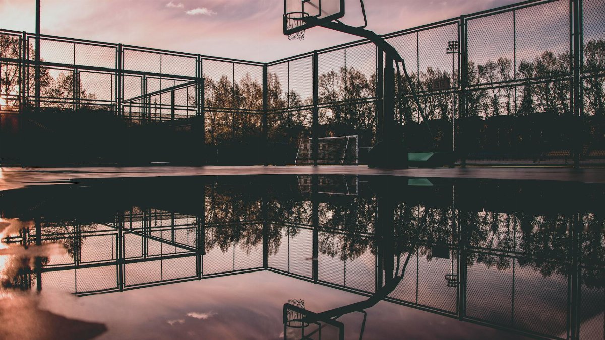 A serene sunset casts reflections on a wet basketball court, creating a tranquil urban scene.